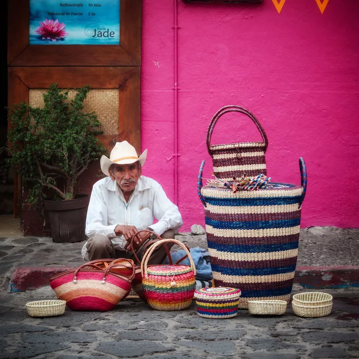 Artesano sentado con sus canastas tejidas frente a una pared rosa mexicano